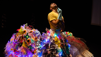 Dark-skinned male dancer with yellow shit and large poofed skirt with lights and many colors.
